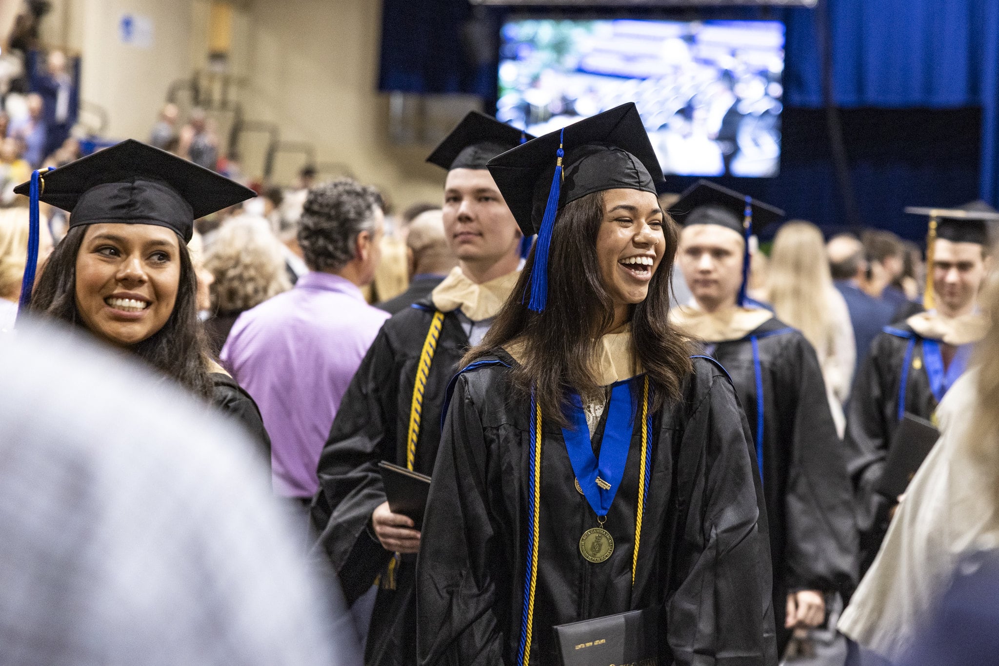 Smiling graduates in caps and gowns at Crummer graduation ceremony, person in foreground wearing honor cords and medal