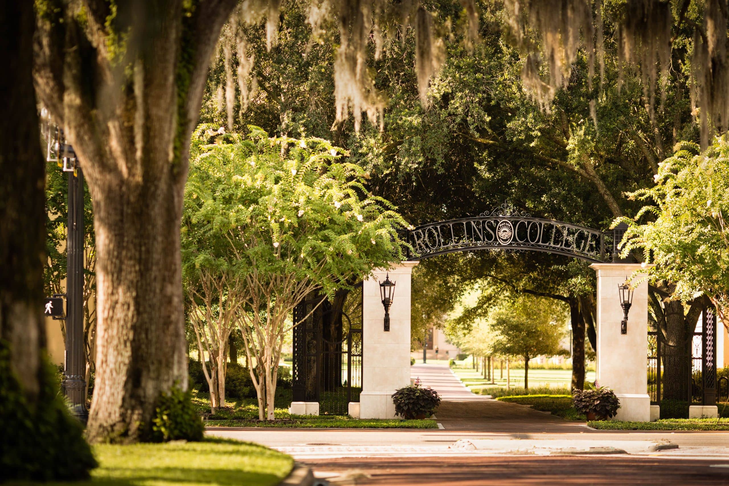 Wrought iron Rollins College arch over brick walking pathway entrance to campus, brick road and lush trees visible