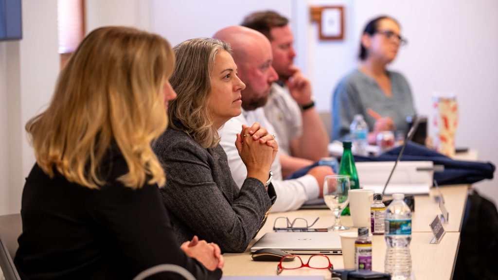A panel of three people, including a woman in the foreground, seated at a long table during a focused professional or academic meeting.